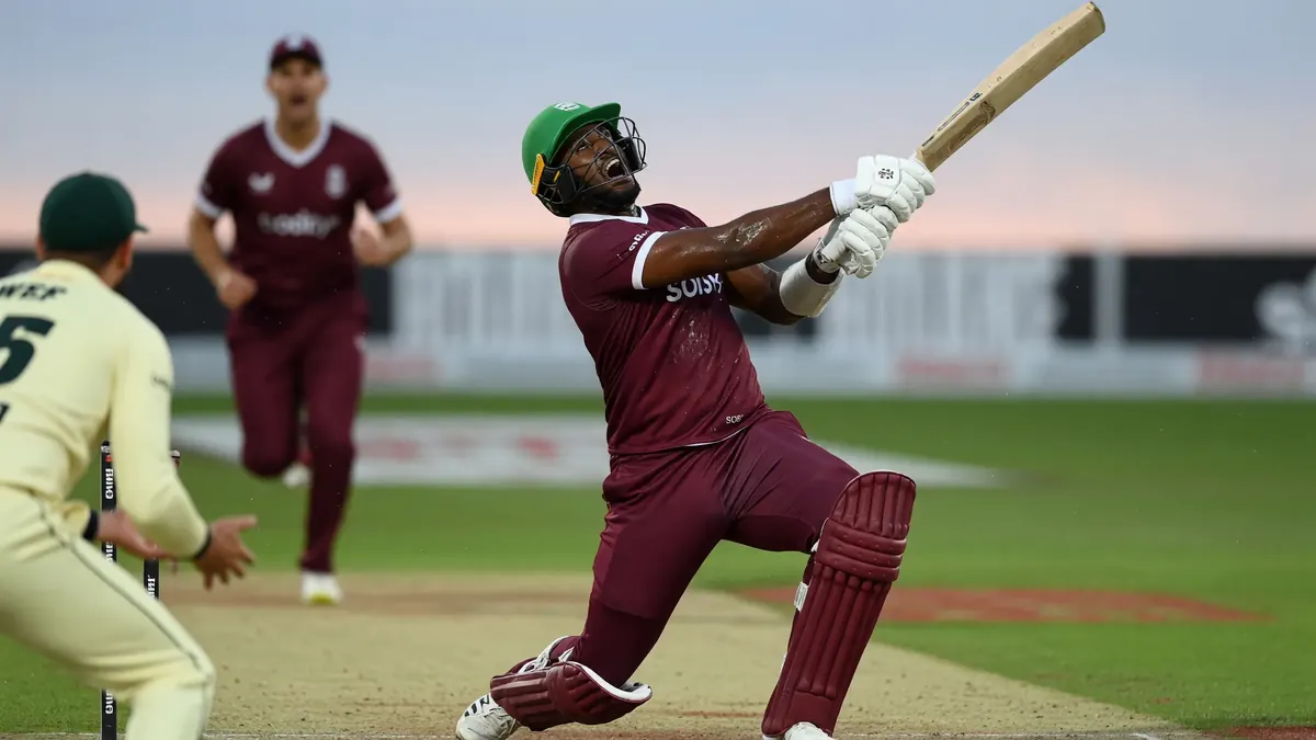 Batter in a maroon kit and green helmet swinging hard during a night cricket match as fielders react behind him.